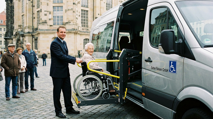 Ein professioneller Fahrer bedient eine sichere hydraulische Hebebühne an einem Kleinbus, um eine ältere Dame im Rollstuhl behutsam in das Fahrzeug zu heben.