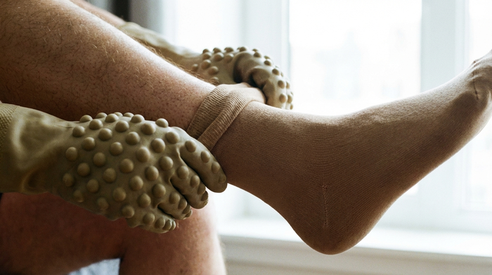 Nahaufnahme von Händen, die spezielle Gummihandschuhe mit Noppen tragen, um vorsichtig einen Kompressionsstrumpf über das Bein zu ziehen. Helles Licht, realistische Szene ohne Text.