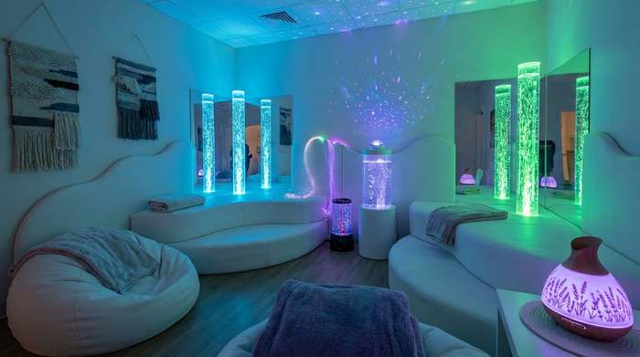Ein gemütliches Snoezelen-Raum-Setup in einer Pflegeeinrichtung. Sanftes, farbiges Licht, bequeme weiße Polsterlandschaften und beleuchtete Wassersäulen mit aufsteigenden Bläschen. Eine ruhige, entspannende Atmosphäre für die Sinne, ohne Menschen.