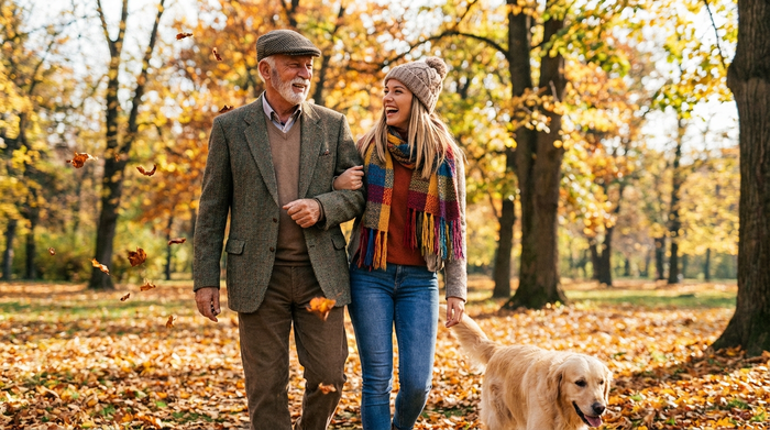 Ein älterer Herr und eine junge, sympathische Alltagsbegleiterin gehen gemeinsam im herbstlichen Park spazieren. Die Blätter sind bunt, beide lachen und genießen die frische Luft in entspannter Atmosphäre.