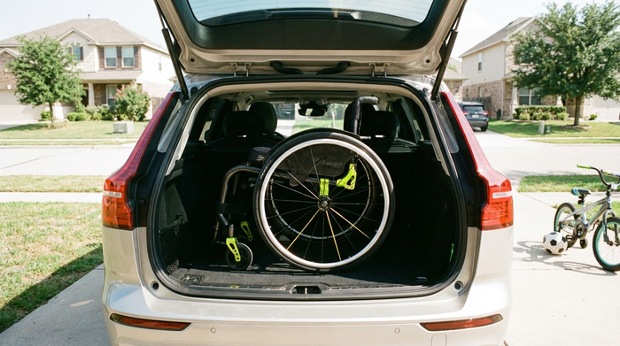 Ein moderner, leichter Rollstuhl steht ordentlich zusammengeklappt im Kofferraum eines Familienautos. Eine helle, saubere Vorstadtszene bei Tageslicht, die Mobilität und Unabhängigkeit ausstrahlt.