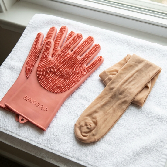 Ein Paar spezielle Noppenhandschuhe aus weichem Gummi liegen ordentlich neben einem frisch gewaschenen, hautfarbenen Strumpf auf einem sauberen weißen Handtuch.