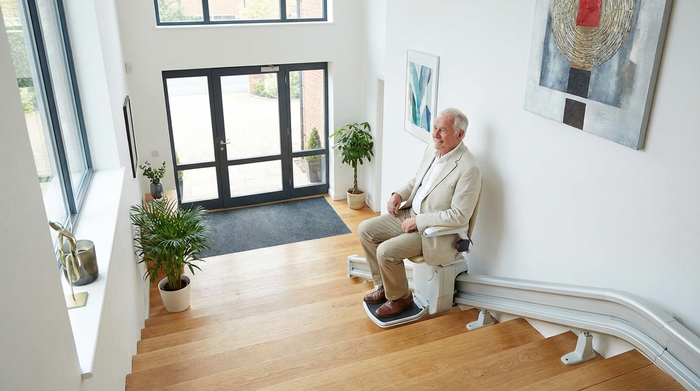 Ein moderner Treppenlift in einem gepflegten, hellen Treppenhaus eines Mehrfamilienhauses. Ein älterer Herr fährt entspannt sitzend die Treppe hinunter in Richtung Haustür.