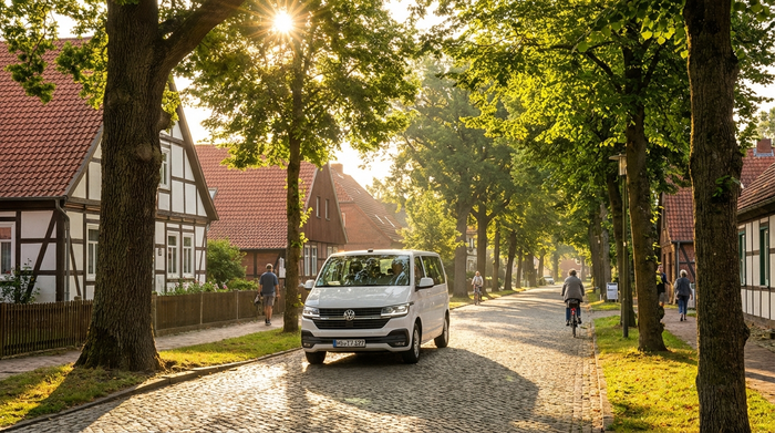 Ein moderner, weißer Kleinbus fährt durch eine von Bäumen gesäumte, ruhige Wohnstraße in einer deutschen Kleinstadt im morgendlichen Sonnenlicht.
