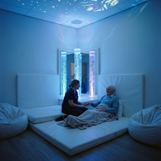 Ein Snoezelen-Raum mit sanfter, bläulicher Beleuchtung, bequemen weißen Liegematten und beruhigend blubbernden Wassersäulen. Eine Pflegekraft sitzt ruhig neben einem sichtlich entspannten Senior.