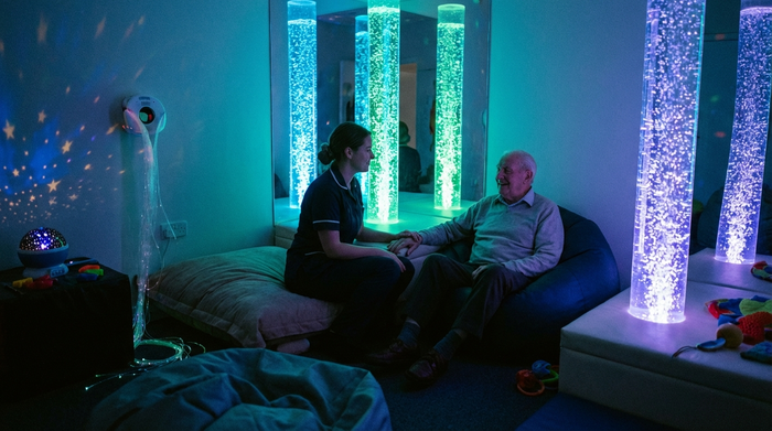 Ein entspannender Snoezelen-Raum mit gedimmtem Licht. Sanft leuchtende, farbige Wassersäulen und bequeme Sitzkissen schaffen eine beruhigende Atmosphäre. Eine Pflegekraft sitzt ruhig neben einem sichtlich entspannten Senior.