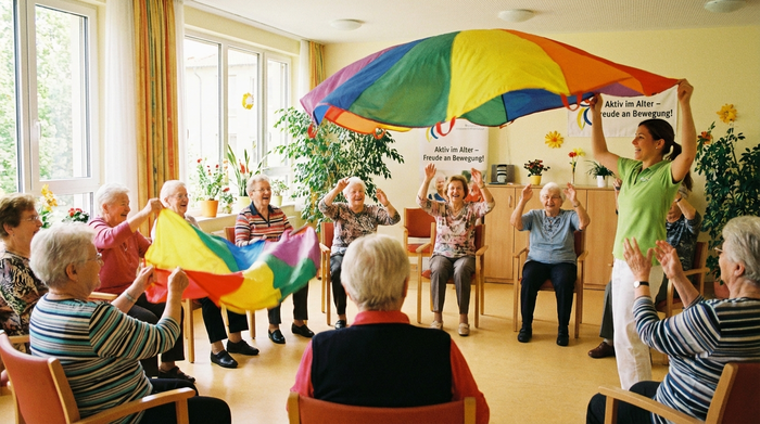 Eine Gruppe von Senioren sitzt im Kreis in einem hellen Raum und macht gemeinsam leichte Sitzgymnastik mit bunten Schwungtüchern. Eine motivierte Therapeutin macht die Übungen vor. Alle lächeln und haben sichtlich Spaß an der Bewegung. Aktive und lebendige Atmosphäre.