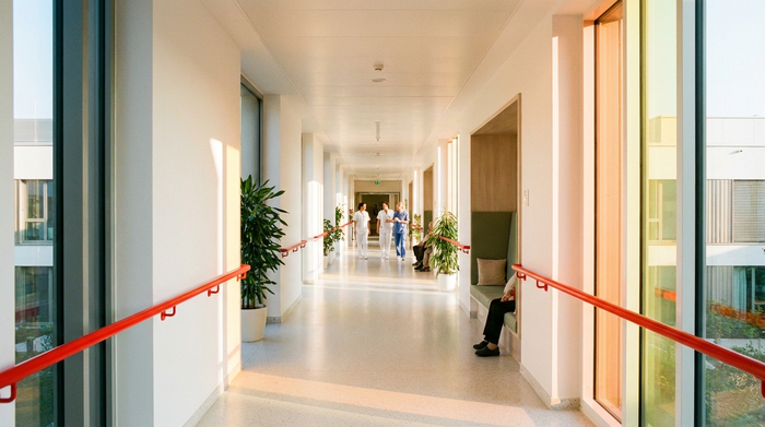 Ein heller, endloser Flur in einer modernen Pflegeeinrichtung ohne dunkle Fußmatten. Handläufe in leuchtendem Rot bieten starken Kontrast. Große Fenster lassen natürliches, biodynamisches Licht herein, das den Raum freundlich erhellt.