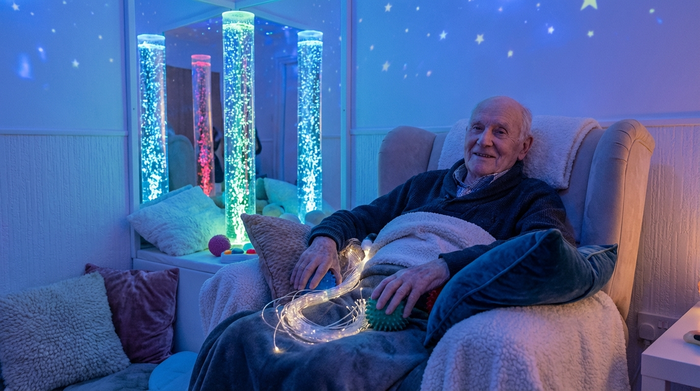 Ein gemütlicher Snoezelen-Raum mit sanfter, bläulicher Beleuchtung und beruhigenden Wassersäulen. Ein älterer Herr sitzt entspannt in einem bequemen Sessel, umgeben von weichen Kissen und taktilen Elementen, die Geborgenheit ausstrahlen.
