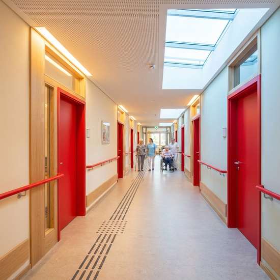 Ein heller, barrierefreier Flur in einer Pflegeeinrichtung mit farblich stark abgesetzten, roten Türen und Handläufen zur besseren Orientierung. Biodynamisches Licht durchflutet den Gang.