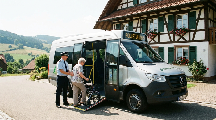 Ein komfortabler, moderner Rollstuhlbus steht mit geöffneten Türen vor einem gepflegten Einfamilienhaus im Schwarzwald. Ein freundlicher Fahrer hilft einer älteren Dame behutsam beim Einsteigen. Sonniges Wetter, realistische Alltagsszene.