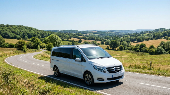 Ein moderner, weißer Kleinbus fährt an einem sonnigen Tag über eine Landstraße. Im Hintergrund sind grüne Bäume und eine weite Landschaft zu sehen, klare und realistische Szene.