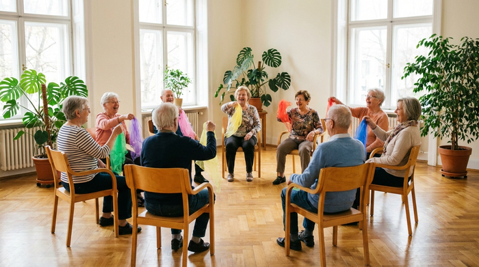 Ältere Menschen sitzen in einem Stuhlkreis und machen leichte Gymnastikübungen mit bunten Chiffontüchern. Ein heller Raum mit großen Fenstern, Zimmerpflanzen und warmem Holzboden. Realistische, fröhliche Atmosphäre.