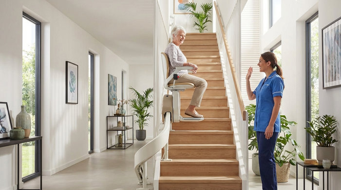 Ein moderner Treppenlift in einem hellen, gepflegten Einfamilienhaus. Eine ältere Person fährt sicher und entspannt die Treppe hinauf, während eine Assistenzkraft am Fuß der Treppe freundlich lächelt.