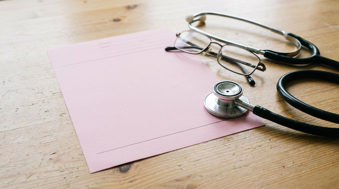 Ein rosa Rezeptformular liegt auf einem hellen Holztisch neben einem Stethoskop und einer Lesebrille. Realistische Nahaufnahme, saubere Umgebung, weiches Tageslicht, keine lesbaren Texte.