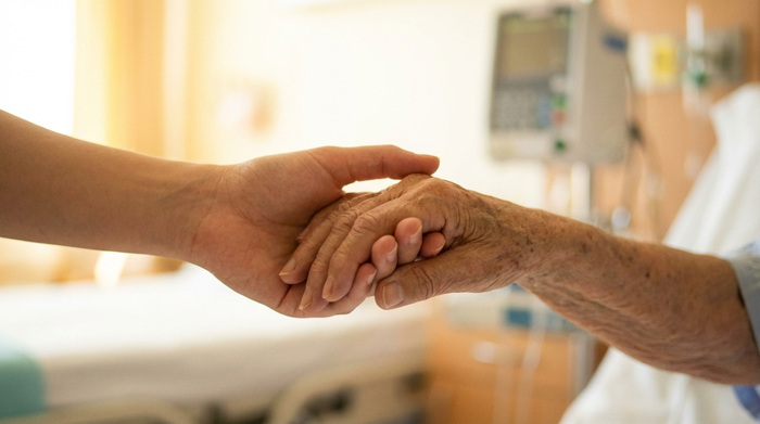 Zwei Hände, eine jung, eine alt, die sich tröstend halten. Im Hintergrund ein unscharfes, helles Krankenzimmer. Sanftes Licht, das Geborgenheit und familiären Zusammenhalt in einer schwierigen Situation ausstrahlt.