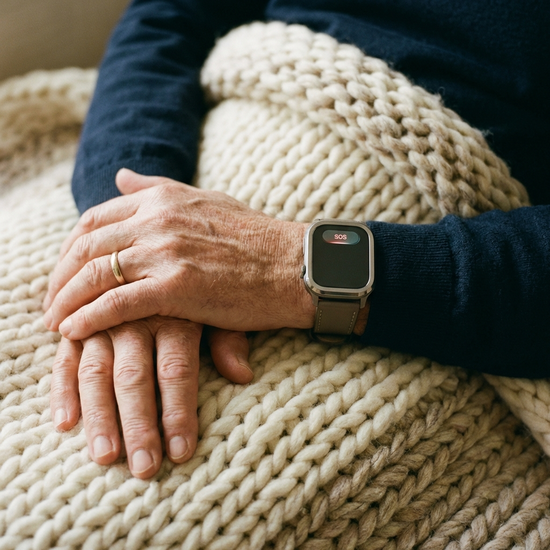 Ein eleganter Hausnotruf-Knopf, der wie eine moderne Armbanduhr aussieht, getragen am Handgelenk eines Senioren. Die Hände ruhen entspannt auf einer weichen Wolldecke.