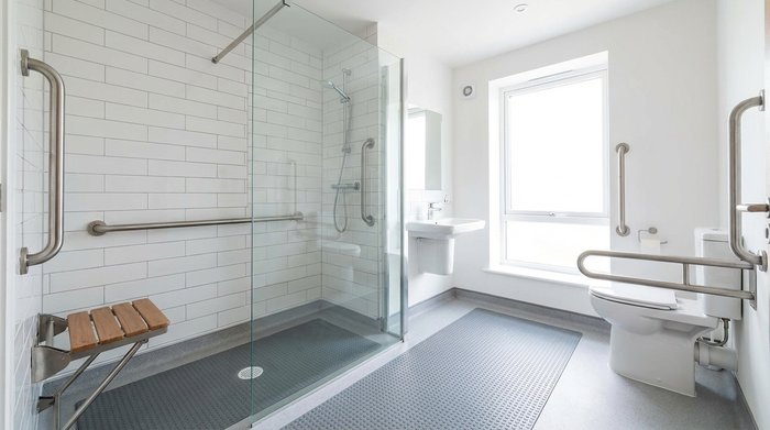 Ein helles, barrierefreies Badezimmer mit einer modernen, ebenerdigen Dusche und stabilen Haltegriffen an den Wänden. Saubere weiße Fliesen, eine rutschfeste Matte auf dem Boden und ein klappbarer Duschsitz. Funktionales, sicheres Design.