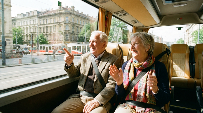 Zwei Senioren sitzen gut gelaunt und sicher angeschnallt in einem hellen Kleinbus. Sie unterhalten sich angeregt und blicken aus dem Fenster auf die vorbeiziehende Stadtlandschaft. Freundliche und fröhliche Stimmung.