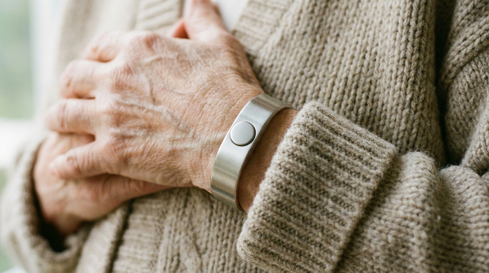 Ein moderner, unauffälliger Hausnotruf-Knopf am Handgelenk einer älteren Person. Nahaufnahme, weiches Licht, fokussiert auf Sicherheit im Alter, gemütlicher Pullover.