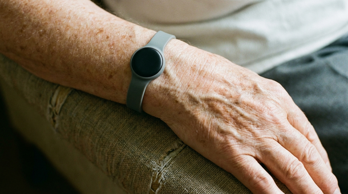 Eine Nahaufnahme eines modernen, unauffälligen Hausnotruf-Armbands am Handgelenk einer älteren Person. Die Hand ruht entspannt auf einer Armlehne. Weiches Licht, fokussiertes Detail, ohne Beschriftungen.