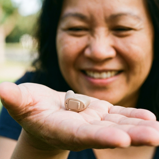 Ein unauffälliges, modernes Hörgerät liegt auf einer Handfläche. Weicher, unscharfer Hintergrund mit einem lächelnden Gesicht.