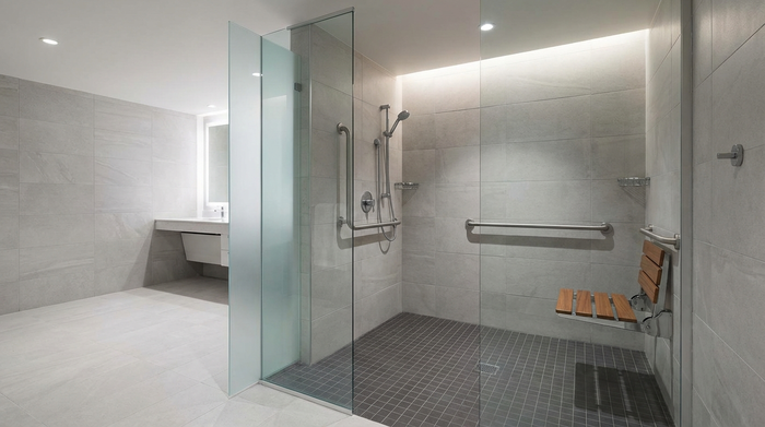 Ein modernes, komplett barrierefreies Badezimmer mit einer großzügigen, bodengleichen Dusche. Die Dusche verfügt über edle Glaswände und einen rutschfesten, dunkelgrauen Fliesenboden. Ein eleganter, hochgeklappter Duschsitz ist an der Wand montiert.