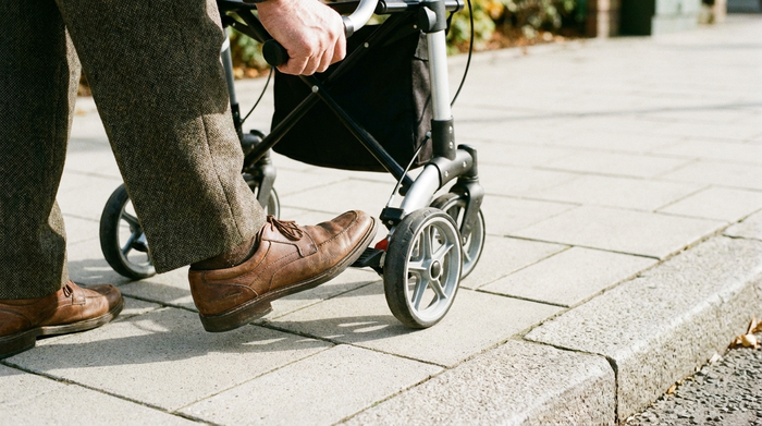 Ein älterer Herr nutzt die Ankipphilfe seines Rollators an einer niedrigen Bordsteinkante. Er drückt sanft mit der Fußspitze auf das kleine Pedal am Hinterrad. Fokus auf Fuß und Hinterrad auf einem sauberen Gehweg.