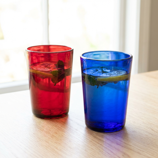 Zwei farbige Trinkgläser, eines rot und eines blau, gefüllt mit frischem Wasser, stehen auf einem hellen Küchentisch. Klarer Fokus, helle und freundliche Beleuchtung.