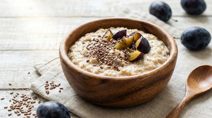 Eine Holzschüssel gefüllt mit cremigem Haferbrei, garniert mit Leinsamen und frischen Pflaumenstücken auf einem hellen Tisch.