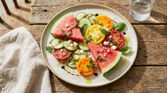 Ein appetitlich angerichteter Teller mit frischen Wassermelonenstücken, Gurkenscheiben und Tomaten auf einem rustikalen Holztisch in hellem, freundlichem Licht.