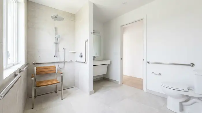Ein helles, modern umgebautes Badezimmer mit einer bodengleichen, geräumigen Dusche. Ein stabiler Duschstuhl steht bereit. Haltegriffe an den Wänden sorgen für zusätzliche Sicherheit. Alles wirkt sauber und barrierefrei.
