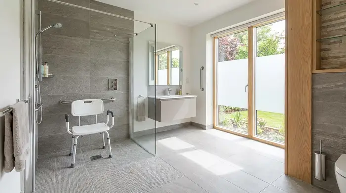 Ein modernes, helles und barrierefreies Badezimmer mit einer bodengleichen Dusche. In der Dusche steht ein sicherer, weißer Duschstuhl mit Armlehnen auf rutschfesten Fliesen. Natürliches Licht fällt durch ein großes Fenster.