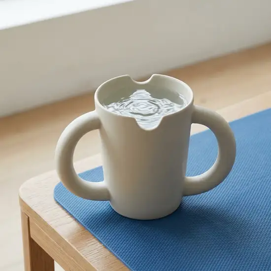 Ein eleganter, moderner Trinkbecher mit zwei großen, geschwungenen Henkeln und einer unauffälligen Nasenaussparung, gefüllt mit Wasser, steht auf einer rutschfesten blauen Silikonmatte.