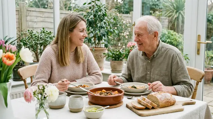 Eine fürsorgliche Tochter sitzt gemeinsam mit ihrem älteren Vater an einem schön gedeckten Tisch. Beide lächeln entspannt, während sie ohne Hektik und in ruhiger Atmosphäre ihr Mittagessen genießen.