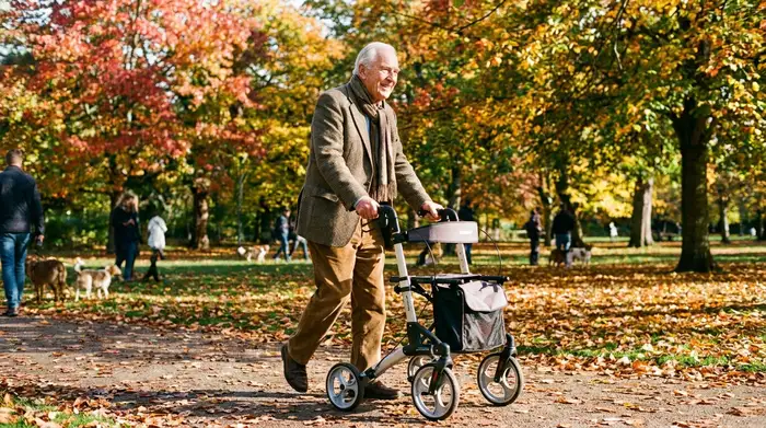 Ein älterer Herr spaziert an einem sonnigen Herbsttag mit einem modernen, leichten Aluminium-Rollator sicher und aufrecht durch einen bunten Park.