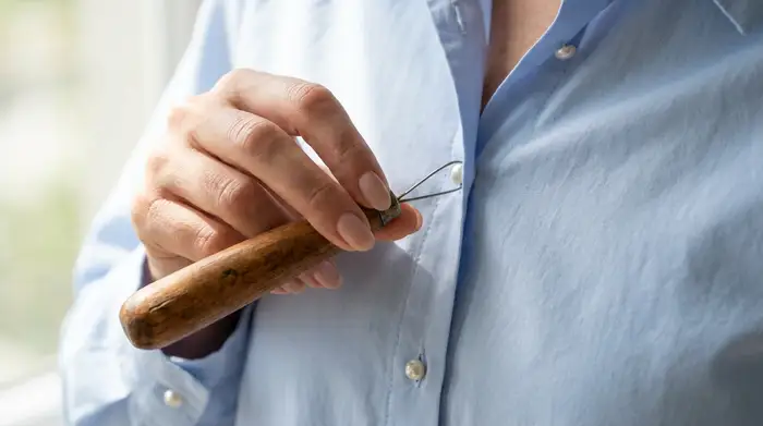 Eine gepflegte Hand benutzt eine spezielle Knöpfhilfe mit einem dicken Holzgriff, um mühelos den kleinen Knopf einer hellblauen Bluse zu schließen. Der Fokus liegt auf der sanften, präzisen Bewegung ohne sichtbare Anstrengung.