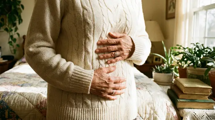 Eine Nahaufnahme der Hände einer älteren Frau, die sanft auf ihrem Bauch ruhen. Sie trägt einen weichen, hellen Strickpullover in einem gemütlichen, lichtdurchfluteten Schlafzimmer.