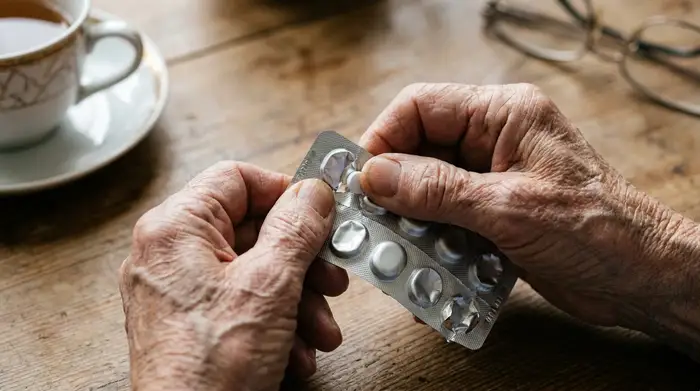Ein detaillierter Blick auf zittrige, faltige Hände eines Seniors, die vorsichtig versuchen, eine winzige weiße Tablette aus einer unhandlichen Blisterverpackung zu drücken.