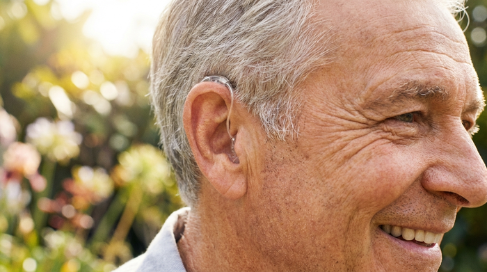 Eine Nahaufnahme eines modernen, fast unsichtbaren Hinter-dem-Ohr-Hörgeräts am Ohr eines lächelnden Seniors. Helle, freundliche Umgebung, hohe Detailtreue.
