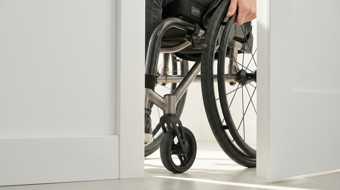 Nahaufnahme eines modernen Rollstuhls, der problemlos durch einen breiten, weißen Türrahmen navigiert. Helles Tageslicht, fokussiert auf den Abstand zwischen Rad und Türzarge. Realistisch, keine Texte.