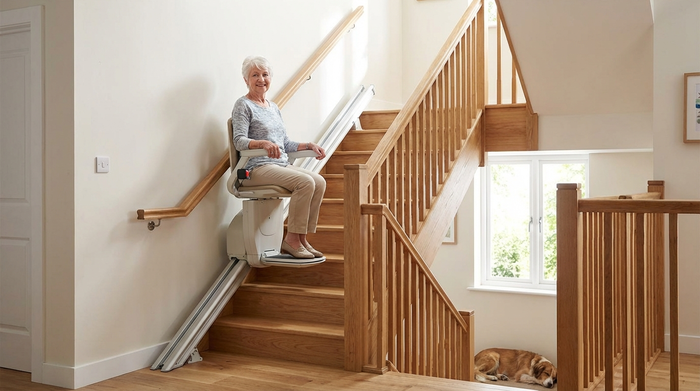 Eine gepflegte Holztreppe in einem hellen Einfamilienhaus. Ein moderner, unauffälliger Treppenlift ist an der Wandseite installiert. Eine rüstige Rentnerin sitzt sicher im Lift und fährt entspannt lächelnd in das obere Stockwerk. Die Treppe hat beidseitig stabile Handläufe.