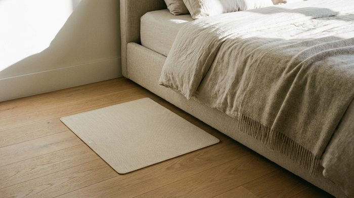 Eine Nahaufnahme einer modernen, flachen Sensormatte auf einem hellen Holzboden neben einem gemütlichen Bett. Die Matte ist dezent und fügt sich harmonisch in das saubere Schlafzimmer ein.