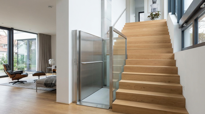 Ein moderner Plattformlift an einer breiten, geraden Treppe in einem gepflegten Einfamilienhaus. Die Plattform ist hochgeklappt und fügt sich unauffällig in das moderne Wohnambiente ein.