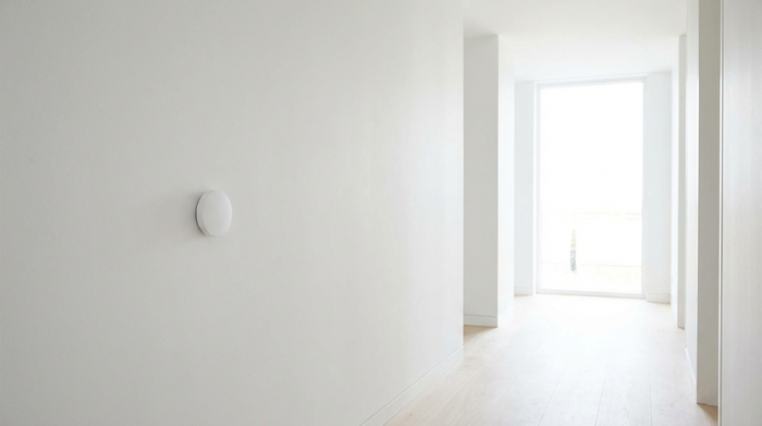 Ein moderner, unscheinbarer Sensor an einer weißen Wand in einem aufgeräumten Flur. Helles Tageslicht, minimalistisches Design, keine störenden Elemente.
