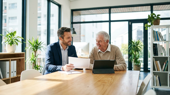 Ein freundlicher, professioneller Pflegeberater sitzt mit einem älteren Herrn an einem Tisch und erklärt ihm geduldig etwas. Beide wirken entspannt und positiv. Helles Büro, realistische Szene, keine Texte.