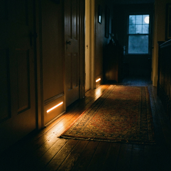Ein dunkler Flur in einem Wohnhaus bei Nacht. Eine kleine, unauffällige LED-Leiste an der Fußleiste wirft ein sanftes, bernsteinfarbenes Licht auf den Holzboden und macht eine Teppichkante deutlich sichtbar.