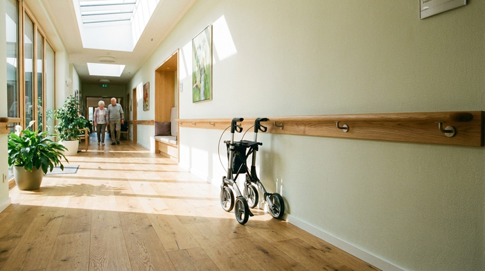 Ein gut ausgeleuchteter Flur in einem modernen Seniorenheim mit einem sicheren Handlauf an der hellen Wand und einem völlig rutschfesten, sauberen Holzboden. Ein Rollator steht ordentlich an der Seite.