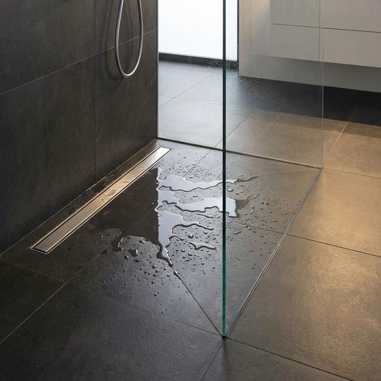 Eine Nahaufnahme einer modernen, bodengleichen Dusche mit eleganter Edelstahl-Duschrinne. Auf dem rutschfesten anthrazitfarbenen Fliesenboden steht das Wasser, das sicher abfließt. Eine gläserne Duschwand im Walk-In-Design rahmt den Bereich ein.
