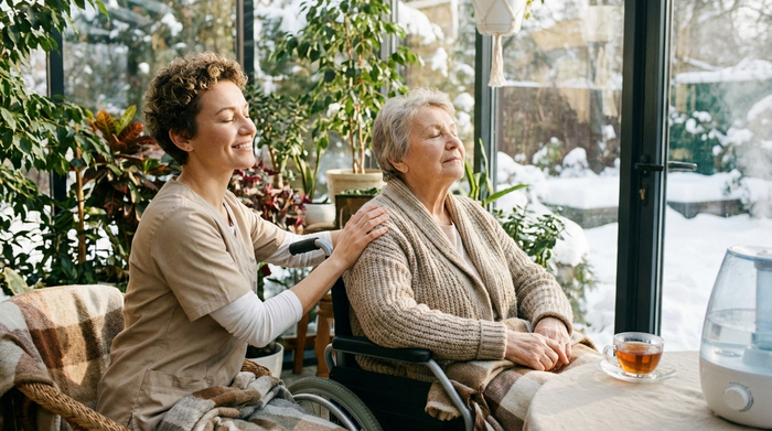Eine freundliche Pflegekraft legt beruhigend eine Hand auf den Rücken einer älteren Dame im Rollstuhl, während sie gemeinsam im hellen Wintergarten eine Atemübung durchführen.